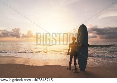 Man Hold Surfboard Standing At Tropical Sunset Beach Background. Summer Vacation And Sport Adventrue