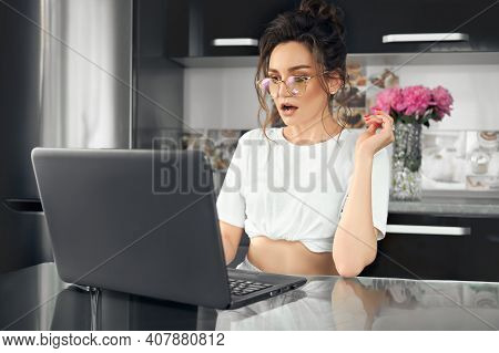 Portrait Of A Young Shocked Woman In Glasses Sitting In Front Of A Laptop Computer, Looking At The S