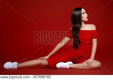 Sport chic style. Beautiful brunette girl posing at studio in red dress and white sneakers. Red background. 