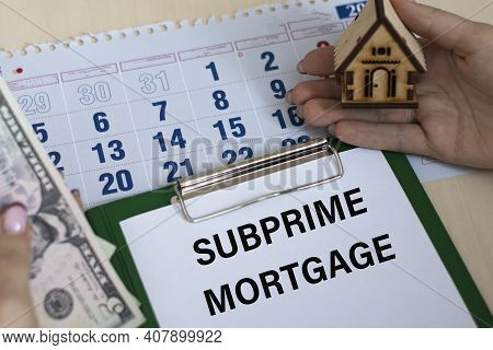 Subprime Mortgage - Word In A Folder Against The Background Of Women's Hands With A House And Money.