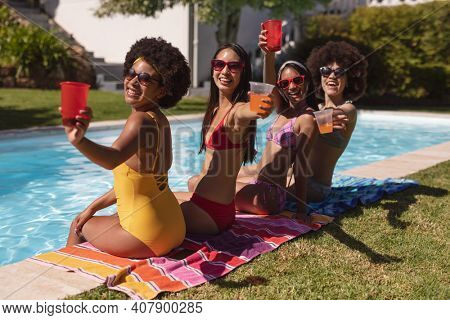 Diverse group of female friends drinking drinks sitting at the poolside. Hanging out and relaxing outdoors in summer.