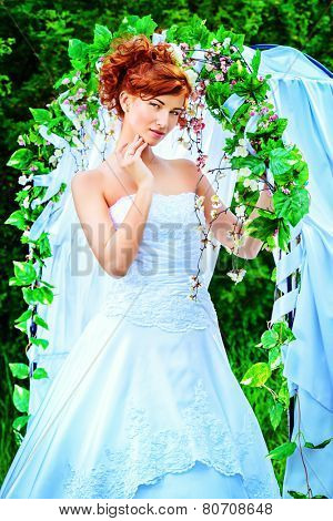 Beautiful bride with chaming red hair stands under the wedding arch. Wedding dress and accessories. Wedding decoration. 