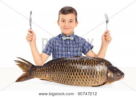 Little kid holding a fork and a knife seated at a table with a huge raw fish in front of him isolated on white background