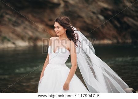 Beautiful luxury young bride in long white wedding dress and veil standing near river with mountains
