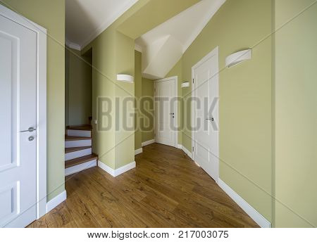 Hall in a modern style with light walls and a parquet on the floor. There are few white doors and a stair with wooden rungs. On the walls there are hanging lamps. Horizontal.