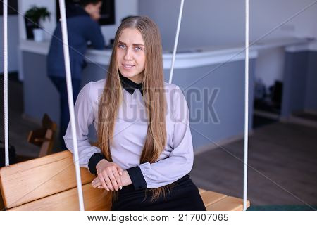 Portrait of cute long-haired successful secretary girl who poses on camera after end of day and smiles in office in daytime. Woman with fair-haired hair dressed in light blouse with black cuffs and skirt below knees of black color. Concept of young and su