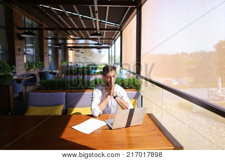 Inventory strategist solving problems and using laptop at cafe table. Clever hardworking man dressed in white shirt working with papers. Concept of  analyzing model forecast  to design contingency plans and catalog displays to make successful, consumer re