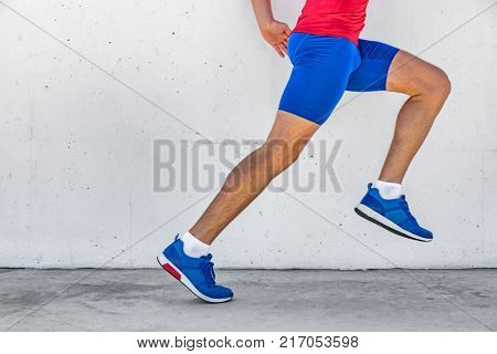 Fitness athlete man running on asphalt sidewalk in city street. Sport lifestyle. Closeup of lower body runner's legs on workout marathon run against white wall background. Shoes, compression clothing.