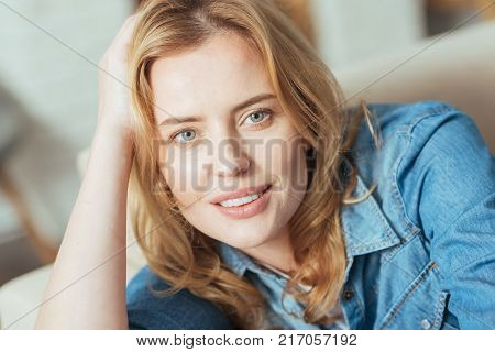 Kind smile. Beautiful calm young woman looking peaceful while resting at home on a comfortable sofa with her hand being on her head