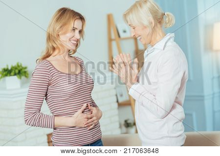 How cute. Pleasant cheerful cute pregnant woman standing and smiling while her emotional positive grandmother looking at her belly