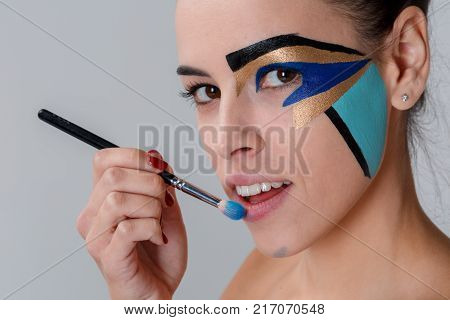Close-up of a brunette, a woman with creative geometric make-up on the face, black lines, holding a make-up brush near her lips, looks into the camera, her mouth slightly open, teeth visible