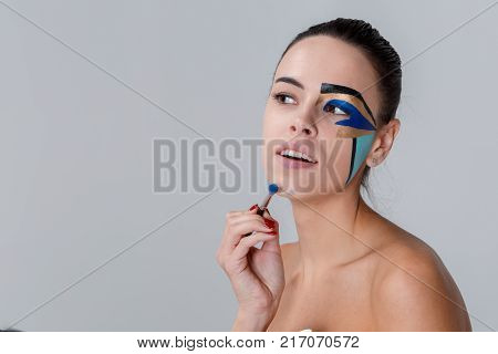 Portrait of a brunette with a bunch on her head, a woman with creative geometric make-up on the face, black lines, holding a make-up brush near the chin, looking away
