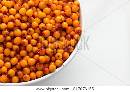 Part of a white plate filled with berries of sea-buckthorn in the bottom left corner on a white background. Isolated top view of a close-up.