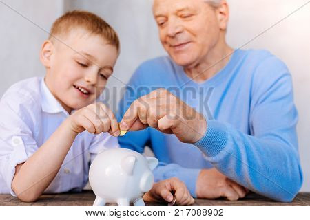 Saving money. Delighted nice pleasant boy sitting together with grandfather and holding a coin while putting it into the money box