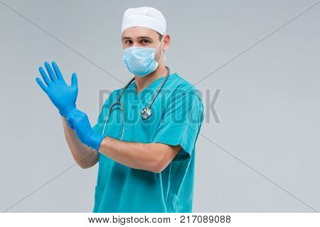 Close-up of a doctor in uniform, in profile, wearing a mask on a gray background, a man in his arms dresses blue, rubber gloves, on his neck hanging strobo, looking at the camera.