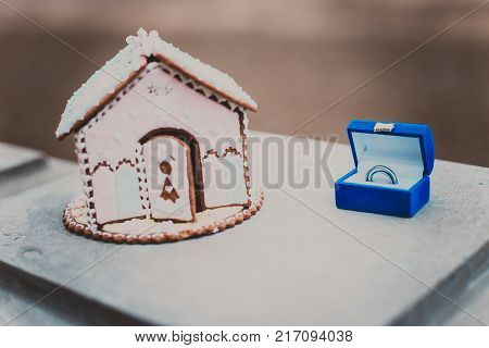 Gingerbread house and two silver wedding rings in blue case on white wooden background.
