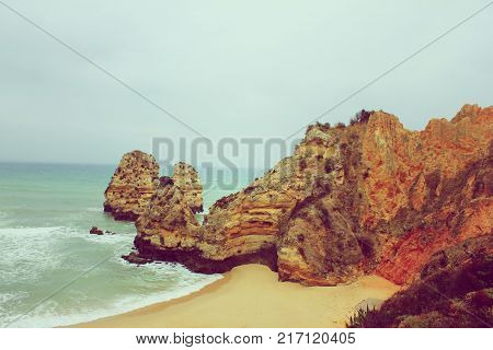 Camilo Beach in Lagos, Algarve Coast, Portugal Vintage Foggy Landscape Wallpaper. Panoramic Ocean View with Stone Cliff Rocks at Sandy Lagoon Beach Shore. Cloudy Sky and Gloomy Nature Background Scene