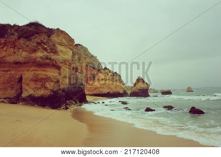 Foggy Landscape of Camilo Beach in Lagos, Algarve Coast, Portugal. Vintage Wallpaper, Panoramic Ocean View with Cliff Rocks and Waves at Sandy Beach Shore. Cloudy Sky and Gloomy Nature Background.