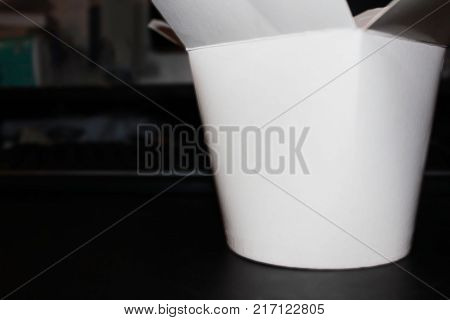 Take Out Delivery Box for Chinese Food on Blurred Office Desk Dark Background. Take Away White Paper Recycled Box with Empty Copy Space in Front of Workplace and Computer Keyboard During Lunch Break.