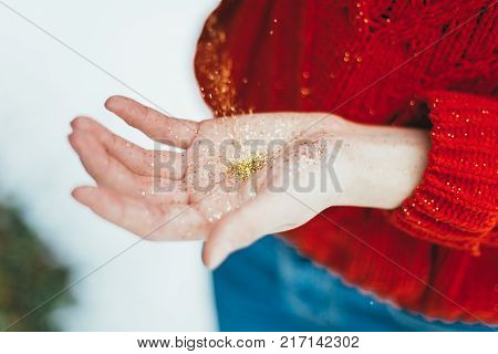 Young curly girl in crystal sunglasses blowing on hands with golden glitters on blue background.