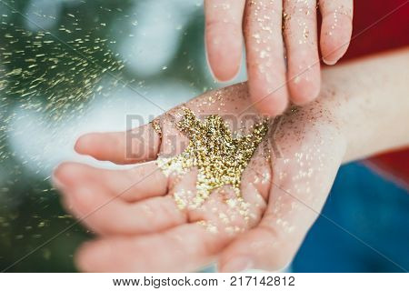Young curly girl in crystal sunglasses blowing on hands with golden glitters on blue background.