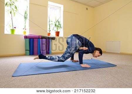 Sportswoman focused on stretch before Pilates class. Girl doing warm up for legs on mat. Female has hair pulled back, concentrated look. Athlete dressed in dark top, yoga legging trains in fitness room. Concept of online training, customizable workouts.