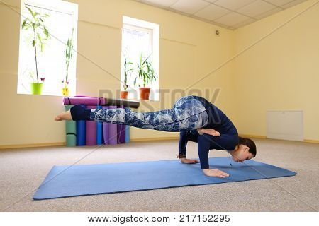 Sportswoman focused on stretch before Pilates class. Girl doing warm up for legs on mat. Female has hair pulled back, concentrated look. Athlete dressed in dark top, yoga legging trains in fitness room. Concept of online training, customizable workouts.