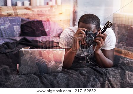Professional device. Handsome nice talented man lying on the bed and using his camera while taking photos