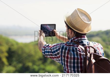 The guy in the hat and with a backpack for traveling makes a phography of a beautiful landscape. He travels to different countries and cities. Perd him a beautiful green maid.