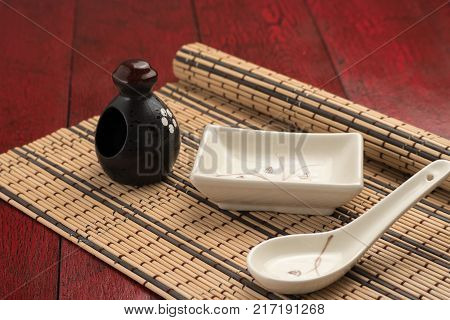 Black porcelain dishes, beige soup tureen with a beige spoon on a bamboo mat. Vintage red background. Front view