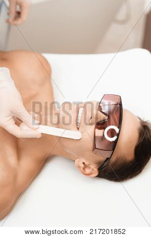 The doctor applies an epilation gel on the man's face. The man laughs, because he is ticklish. He lies on a couch in a modern beauty salon.