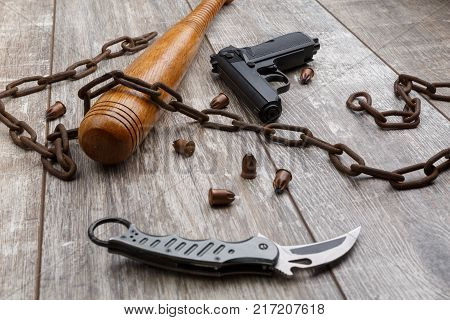 A rusty chain with a bat, a folding knife, bullets and a gun lies on a wooden background. Side view at an angle
