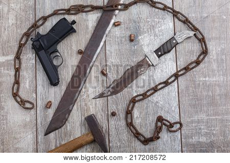 A rusty chain with a rusty homemade machete, a pistol, bullets, a knife and a hammer on a wooden background. View from above