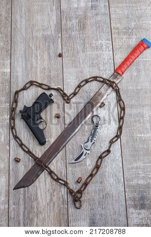A rusty chain in the shape of a heart and a machete that pierces the heart, pistol with a knife and a wooden background. View from above