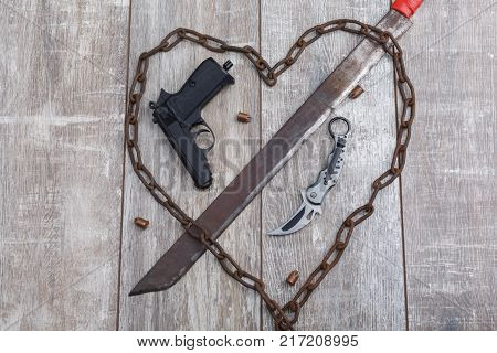 A rusty chain in the shape of a heart and a machete that pierces the heart, pistol with a knife close-up a wooden background. View from above