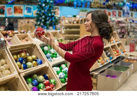 Cheerful happy customer girl choosing gifts for Christmas and New Year. She is taking red balls Christmas tree decorations. difficult decision. Various choice. She is smiling