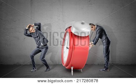 A businessman covers his head form a giant red retro alarm clock being winded up by another businessman. Time bomb. Strict deadline. Teamwork rules.