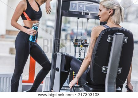 attractive athletic woman working out with gym machine while her partner resting with water bottle next to her at gym