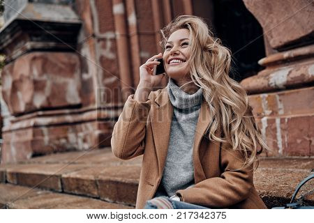 So good to hear you! Beautiful young woman talking on her smart phone and looking away with smile while sitting on stairs outdoors