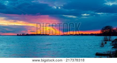 A Winter Sunset on the Lakeshore in Lachine Quebec. The silhouette of a  leafless tree, against  the blue sky and orange clouds on a cold winters afternoon, illuminate the scene.