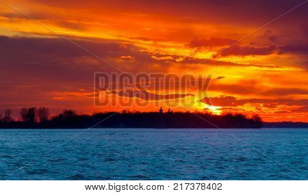A Winter Sunset on the Lakeshore in Lachine Quebec. The silhouette of a  leafless tree, against  the blue sky and orange clouds on a cold winters afternoon, illuminate the scene.