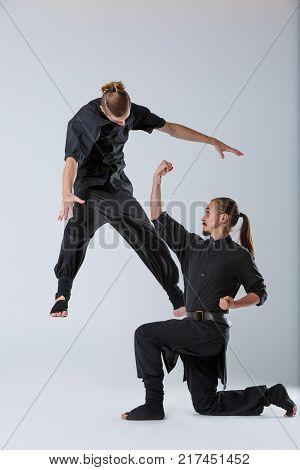 Ninja young, with long hair in black uniform, beats an uppercut standing on one knee of the opponent on a gray background