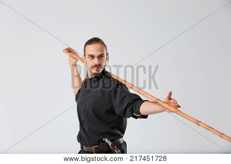 A ninja man young in a black uniform is seriously attuned. It is standing half-turned with a war stick behind the neck and holding hands on a gray background