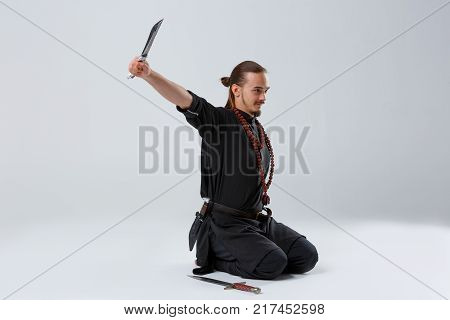 A ninja man with long hair, with a beard in black uniform, kneels with a knife in his outstretched arm on a gray background