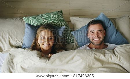 Top view of couple having fun in bed hiding under blanket and looking into camera