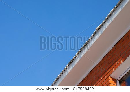 sunny day blue sky. On the roof of the house the snow melts flows and turns into icicles. House of red brick white cornice corner of the white window.