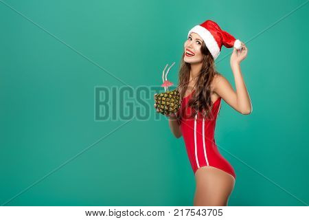 beautiful woman in red bikini and christmas hat drinks summer cocktail in pineapple. christmas holiday