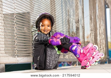 Portrait of beautiful African girl wearing roller skates and protective gear, sitting next to the wire mesh fence at outdoor rollerdrom