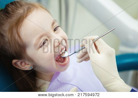 Happy child in comfortable dentist chair with paper napkin on chest and doctor in rubber gloves who checks mouth with sharp metal instrument and magnifying glass.