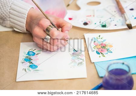 handcrafting, painting, hobby concept. close up of female arm that is holding thin brush and drawing flowers on the heavy stock paper that is rather rough and used for greetin cards